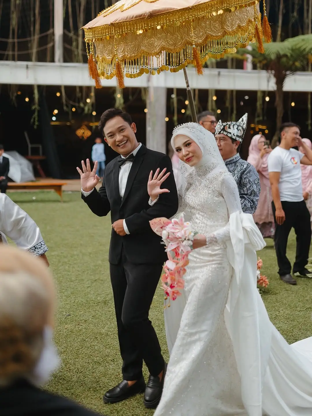 A newly married couple walking across the lawn under a gold ceremonial umbrella as guests look on